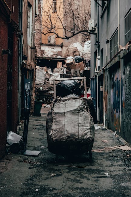 In the foreground, a collection of various discarded household items, including large cardboard boxes, plastic containers, and tangled pieces of wood and metal, all placed on a paved driveway surface.