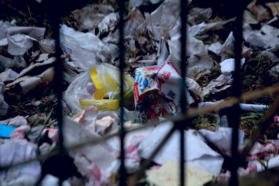 A close-up view of a pile of mixed general waste and rubbish behind black metal fencing. The debris includes crumpled paper, plastic wrappers, and discarded packaging with various textures and colors