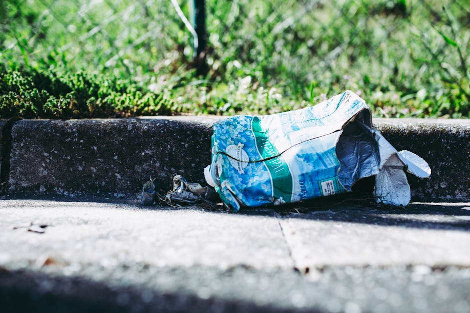 A close-up view of a pile of mixed general waste and rubbish behind black metal fencing. The debris includes crumpled paper, plastic wrappers, and discarded packaging with various textures and colors